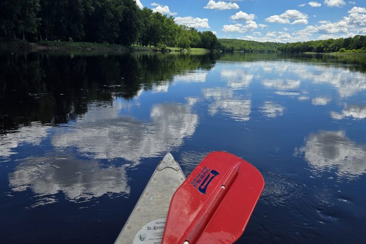 Red canoe paddle rests on a dock by a calm lake reflecting clouds and trees.