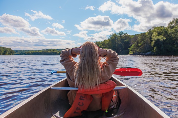 Person with long hair in a canoe on a lake, holding a paddle, surrounded by trees and clouds.